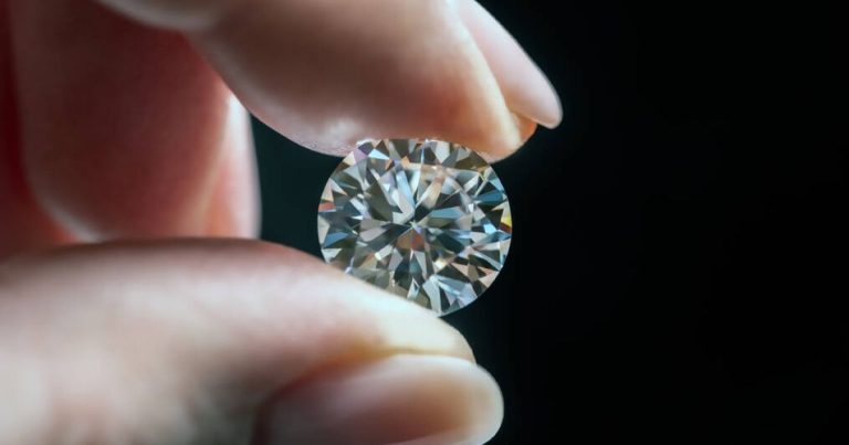 Női kéz ujjai közt egy csiszolt gyémántot tart, fekete háttér előtt